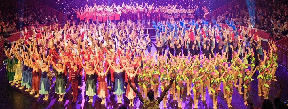 A large ensemble of young dancers on stage with arms raised