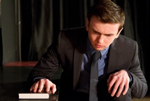 A performer in a suit delivering a dramatic scene at a table